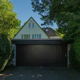 Een huis met een puntdak en een donkere garagepoort, tussen hagen