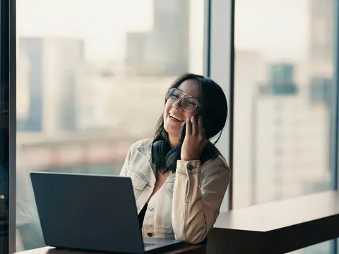 Een vrouw belt lachend bij het raam, met een laptop voor zich