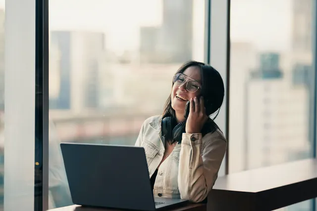 Een vrouw belt lachend bij het raam, met een laptop voor zich
